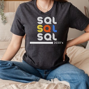 Op de afbeelding: Een donkergrijs T-shirt met een grafisch ontwerp van het woord "SQL" dat drie keer wordt herhaald in verschillende kleuren: geel, oranje en blauw. De tekst "2020's" staat onder de grafische print.