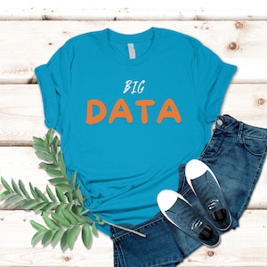 May include: A turquoise blue t-shirt with the text "BIG DATA" in orange letters. The shirt is laid flat on a white wooden surface with a pair of blue jeans and white sneakers.