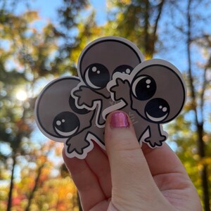 May include: Three gray alien stickers with large black eyes and small arms are held up against a blurred background of trees and sky. The stickers have a white outline and a cartoonish design.