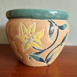 May include: A ceramic plant pot with a tan base and a green rim. The pot features a raised floral design with yellow flowers and green leaves. The design is set against a tan background.