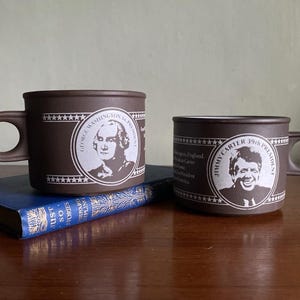 Könnte beinhalten: Zwei braune Tassen mit Henkeln, jeweils mit einem Porträt eines US-Präsidenten. Eine Tasse zeigt George Washington, die andere Jimmy Carter. Die Tassen stehen auf einem blauen Buch.