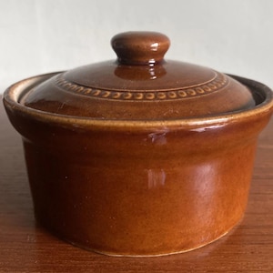 May include: A small, round, brown ceramic lidded container with a glossy finish. The lid features a central knob handle and a decorative embossed beaded border pattern around the edge. The container has straight sides and a smooth, uniform glaze.