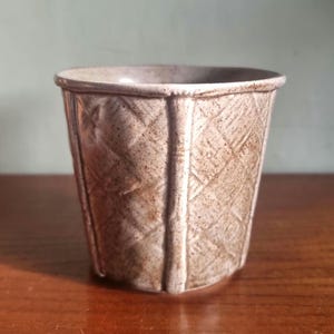 May include: A small, beige ceramic planter with a textured, geometric pattern. The planter has a slightly flared rim and vertical lines that divide the sides into panels. The surface has a speckled appearance.