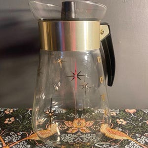 May include: A vintage clear glass jug with a gold-coloured metal band and black handle. The jug features gold starburst and line designs. The jug has a clear plastic spout and a black lid. The jug has measurements marked on the side.
