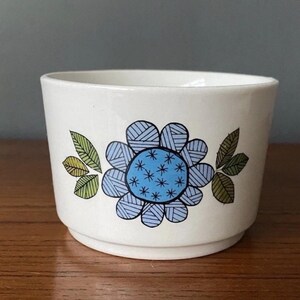 May include: A white ceramic bowl featuring a blue floral design with a light blue centre and green leaves. The bowl is on a wooden surface.