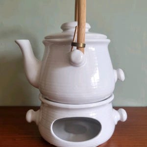 May include: White ceramic teapot with a wooden handle and a matching warmer base. The teapot has a curved spout and a lid. The warmer base has an oval opening. The handle is made of light-coloured wood and is attached with a brass ring.