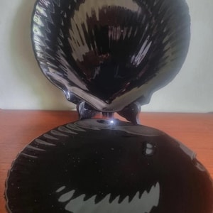 Könnte beinhalten: Zweiteilige schwarze Servierschale in Muschelform. Das Oberteil ist eine tiefe Schüssel, das Unterteil ein flacher Teller. Beide Teile haben einen gewellten Rand und eine glänzende Oberfläche.