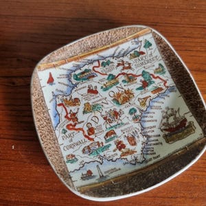 May include: A vintage ceramic plate with a map of Cornwall and Somerset. The plate is square with a textured brown border. The map is detailed with illustrations of landmarks and towns, and the text includes place names.