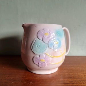 May include: A light pink ceramic jug with a handle and spout. The jug is decorated with embossed flowers in pastel colours of purple, blue, and green, with yellow centres. The jug sits on a wooden surface.