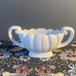 May include: A white ceramic bowl with two curved handles. The bowl has a scalloped rim and sits on a small pedestal base. The bowl is likely used for serving or display.