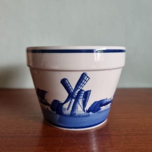 May include: A small, off-white ceramic pot featuring a blue windmill design. The pot has a blue band around the rim and a blue landscape scene around the base. The windmill has four blades and is set against a blue sky.