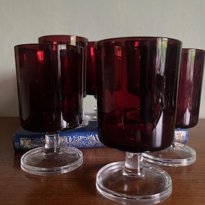 May include: A set of five vintage wine glasses. Each glass has a deep red bowl and a clear, circular base. The glasses are arranged on a wooden surface, with a blue book in the background. The glasses are suitable for serving wine.