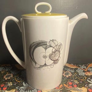May include: White ceramic teapot with a yellow lid and handle. The teapot has a black and white apple illustration. The handle is curved, and the spout is angled. The teapot is likely for serving tea or coffee.