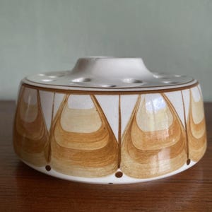 May include: A white ceramic flower frog with a brown and tan teardrop pattern. The frog has a circular base with holes for arranging flowers. The design is mid-century modern, and the colour palette is neutral.