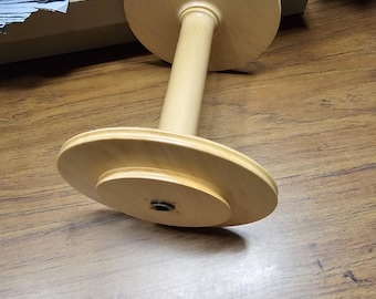 May include: A wooden yarn swift, featuring a central spindle connecting two circular platforms. The swift is made of light-colored wood and is designed to hold yarn for knitting or crocheting. The swift is resting on a wooden surface.