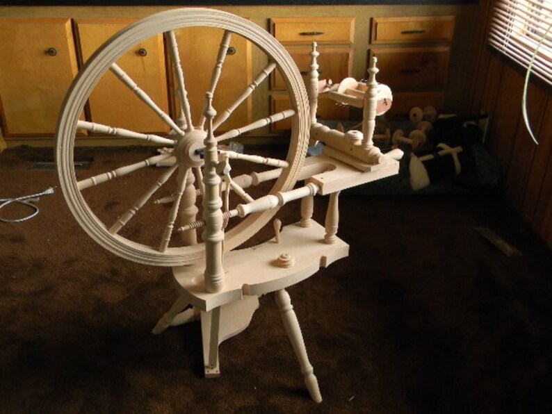 Puede incluir: Una rueca de madera de color claro que presenta una gran rueda circular con m&uacute;ltiples radios, un marco horizontal con patas de soporte torneadas y un mecanismo de hilado situado en la parte trasera. Estructura de madera sin tratar.