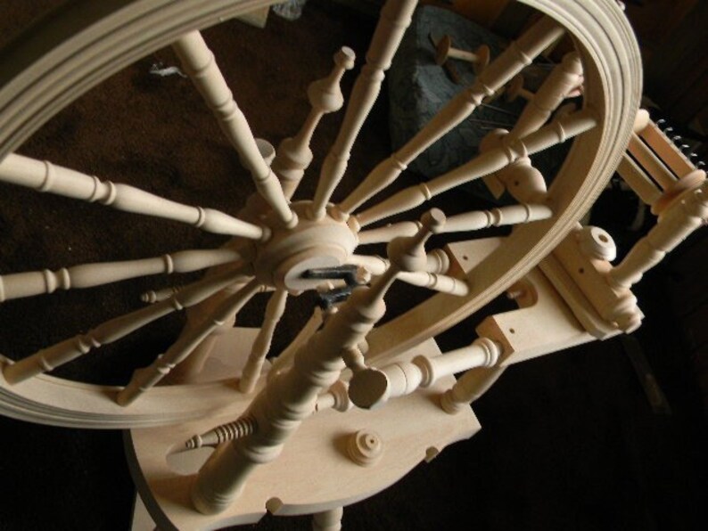 Puede incluir: Una rueca de madera de color claro que presenta una gran rueda circular con numerosos radios torneados, un cubo central y una estructura de base horizontal. El dispositivo incluye un mecanismo de carrete y un volante, todo hecho de madera lisa.