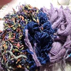 May include: Close-up of a pile of colorful yarn in shades of purple, blue, green, and orange. The yarn appears to be a mix of textures, including thick, chunky strands and finer, more delicate threads. Ideal for fiber art projects.