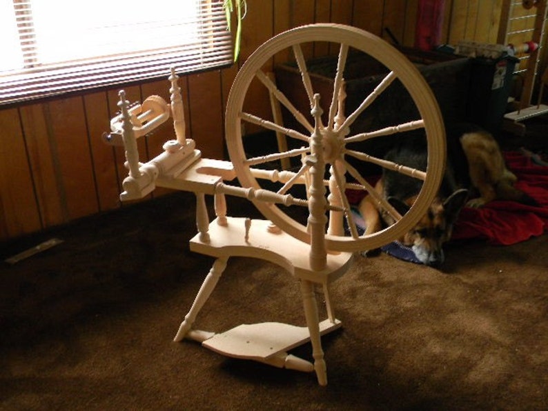 Puede incluir: Una rueca de madera de color claro que presenta una gran rueda motriz circular con m&uacute;ltiples radios, un conjunto de volante, un marco horizontal y un pedal. La estructura est&aacute; hecha de madera lisa sin acabado con detalles de husos torneados.