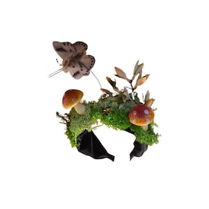 Pode incluir: Uma faixa de cabeça caprichosa com uma borboleta, cogumelos e vegetação. A borboleta tem um padrão salpicado. Os cogumelos têm chapéus castanhos e vermelhos. A faixa de cabeça é preta e coberta de musgo e folhas.