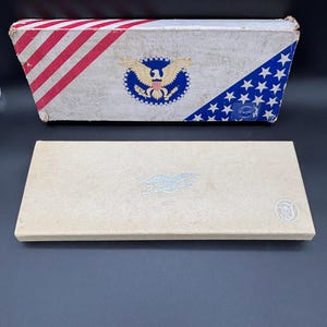 May include: A rectangular beige box with a silver embossed eagle emblem and a circular logo, positioned in front of a larger rectangular box featuring a patriotic design with red and white stripes, a central eagle crest, and a blue field with white stars.