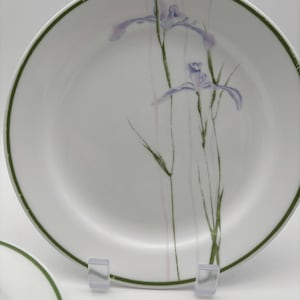 May include: White ceramic plate with a green rim and a watercolor design of purple iris flowers. The plate features a delicate floral pattern with green stems and leaves. The design is centered on the plate, creating an elegant and artistic look.
