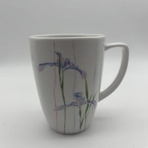 May include: White ceramic mug with a curved handle. The mug features a floral design of blue and purple irises with green stems and vertical pink and blue stripes. The mug is a standard size for coffee or tea.