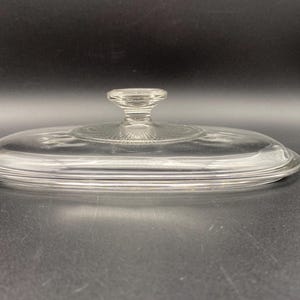 May include: Clear glass oval-shaped lid with a knob on top. The lid has a raised rim around the edge.