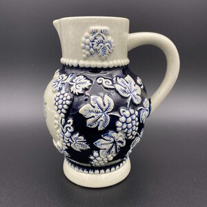 May include: A white ceramic pitcher with a blue and white grape design. The pitcher has a handle and a round body.