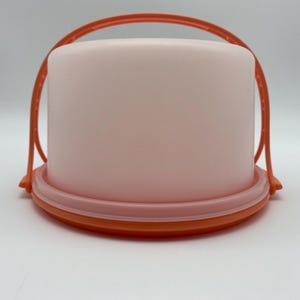 May include: A round, light pink container with an orange handle and base. The container has a cylindrical shape and a lid that fits snugly on the base. The handle arches over the top, providing a convenient way to carry the container.