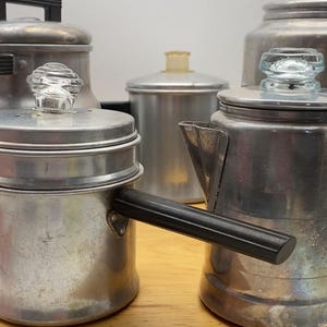 May include: A collection of vintage aluminum kitchenware, including a coffee pot with a black handle, a small pot, and several canisters. The lids feature clear glass knobs. The items are arranged on a wooden surface.