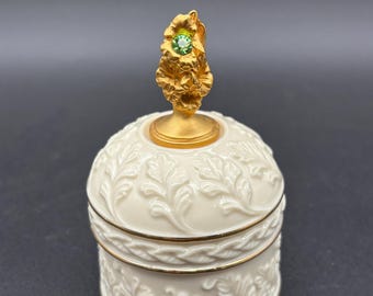 May include: A round, cream-colored porcelain trinket box with embossed leaf patterns and gold trim. The lid features a gold-toned, textured floral finial topped with a single, small, round, light green gemstone set in the center.