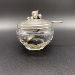 May include: A glass sugar bowl with a pewter lid and a small spoon. The lid is decorated with a cluster of grapes and leaves.