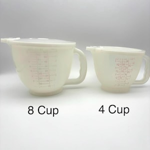 May include: Two off-white measuring cups with handles and lids. The larger cup is labeled "8 Cup" and the smaller cup is labeled "4 Cup". Both cups have red measurement markings on the side.