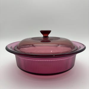 May include: A vintage, round, ruby-colored glass casserole dish with a matching lid. The dish has a ribbed exterior and a clear glass lid with a small, pointed handle. The casserole dish is designed for baking and serving.