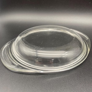 May include: A clear glass lid with two handles. The lid is round and has a slightly domed shape.