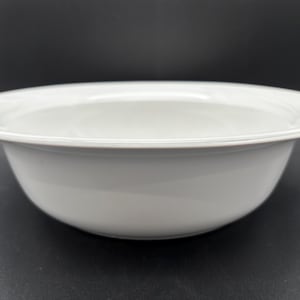 May include: A plain white ceramic bowl with a wide, flat rim. The bowl features a smooth, glossy finish and a rounded base, presenting a clean and simple design. The interior is deep and concave, while the exterior walls curve gently outward.