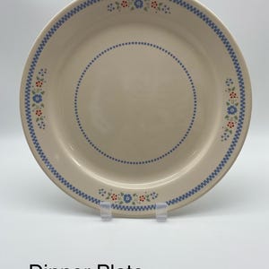 May include: Cream-colored dinner plate with a blue dotted border and a blue zigzag edge. The plate features small floral clusters in blue, red, and green. The text "Dinner Plate" is displayed at the bottom of the image.