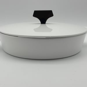 May include: A white oval-shaped casserole dish with a lid. The lid has a black handle. The dish has two small handles on either side. The casserole dish is made of ceramic or enamel and is suitable for cooking and serving.