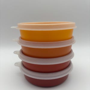 May include: A stack of four colorful food storage bowls with clear lids. The bowls are in shades of orange and red, with the top bowl having a peach-colored lid. The lids have small tabs for easy opening. The bowls are stacked on top of each other.