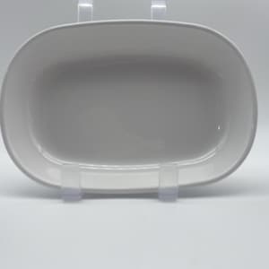 May include: A white, oval-shaped serving dish with a slightly raised rim. The dish is supported by clear plastic stands, and the background is a plain white. The dish appears to be made of ceramic or porcelain.