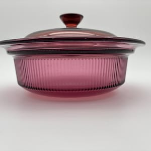 May include: A vintage, round, ruby-colored glass casserole dish with a matching lid. The dish has vertical ribbed detailing and a dark red knob on the lid. The dish is designed for cooking and serving.