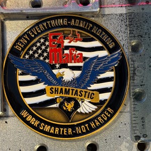 May include: A round, black and gold coin with the text "DENY EVERYTHING - ADMIT NOTHING" and "WORK SMARTER - NOT HARDER". The coin features an eagle with blue wings and the words "E4 Mafia" and "SHAMTASTIC".