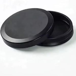 Può includere: Un contenitore nero e rotondo con un coperchio leggermente socchiuso, che rivela l'interno. Il contenitore e il coperchio sono realizzati in materiale nero opaco, su sfondo bianco. Il design è semplice e moderno.