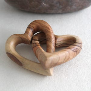 Puede incluir: Dos corazones de madera entrelazados, tallados en madera marrón claro con vetas marrones más oscuras. Los corazones son lisos y pulidos, con un acabado natural de madera.