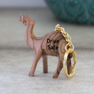 Op de afbeelding: Een houten kameel sleutelhanger met de woorden "Drive Safe" gegraveerd op de zijkant. De kameel is lichtbruin met een goudkleurige ketting en sleutelring. De sleutelhanger is een decoratief accessoire.