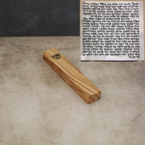 Puede incluir: Estuche de mezuzá de madera con un emblema plateado y texto hebreo. La caja es de madera clara con veta natural. El pergamino es visible en el fondo.