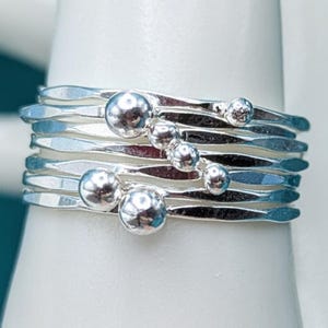 May include: A stack of silver rings with a hammered texture and small, round, polished silver beads. The rings are displayed on a white surface, showcasing their reflective quality and intricate design. The rings are a jewelry item.