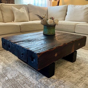 May include: A dark wood coffee table with a rustic, reclaimed look. The table has a thick, rectangular top and four chunky legs. A vase with flowers sits on the table.