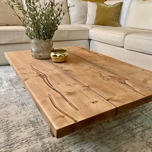May include: A rustic wooden coffee table with a natural wood finish. The table has a rectangular top and is supported by four legs. A small potted plant and a gold bowl are on the table.
